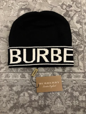Svart mössa från Burberry - Säljer en svart mössa från Burberry med bred vit kant och stor logotyp i vitt runt om. Klassisk och stilren design med ribbstickad känsla och mjukt material. Perfekt accessoar för dig som vill ha något trendigt och exklusivt.