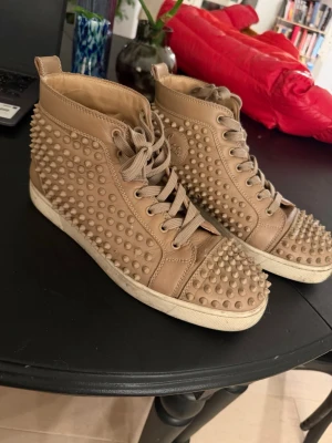 Beige sneakers med nitar - Säljer ett par beige high-top Loubs med nitar över hela skon. Skorna har snörning och en platt vit sula. Tillverkade i skinn. Perfekta för dig som vill sticka ut med din stil. Kommer utan låda, dustbag finns kvar. Tvättar skorna innan frakt. Mvh Grischelino 🙏