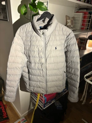 Ljusgrå dunjacka från Polo Ralph Lauren - Snygg ljusgrå dunjacka från Polo Ralph Lauren med diskret logga på bröstet. Jackan har ståkrage, dragkedja framtill och två sidofickor med dragkedja. Quiltad design som ger en modern look och håller värmen under kalla dagar.