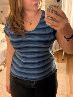 Vintage Blå randig t-shirt - Säljer en söt blå t-shirt med smala ränder i olika blå nyanser. T-shirten har v-ringning, korta ärmar och har en skön passform. Perfekt till jeans eller kjol för en avslappnad look. Tröjan är köpt second hand originalpris 150kr. Prislappen är bortriven så vet inte vart den kommer från originellt.100% bomull