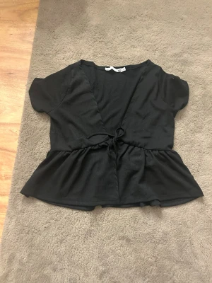 Svart omlottopp med knyt - Svart topp med omlottmodell och knyt i midjan. Kortärmad och med volang nedtill som ger en snygg siluett. Perfekt att styla med jeans eller kjol för en trendig look.