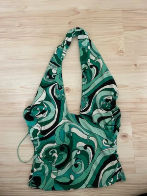 Grön mönstrad halterneck topp - Säljer en snygg halterneck topp med grönt, svart och vitt abstrakt mönster. Toppen har djup v-ringning och knyt i nacken, perfekt för dig som gillar statement-plagg. Materialet känns mjukt och följsamt. Halterneck!