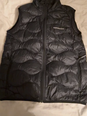 Peak Performance väst - Säljer en svart dunväst från Peak Performance med quiltad design och hög krage. Västen har dragkedja framtill och två sidofickor med dragkedja. Perfekt för lager-på-lager och riktigt snygg till höst- och vinteroutfits.