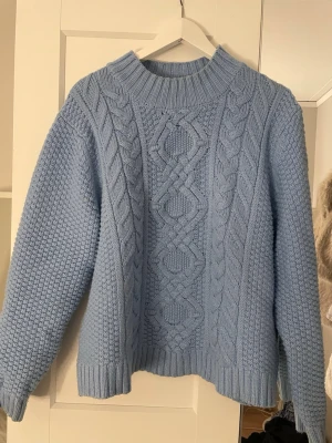 Ljusblå kabelstickad tröja - Mysig ljusblå stickad tröja med grovt kabelmönster framtill och bred ribbad krage. Tröjan har lång ärm och ribbade muddar vid ärmslut och nederkant. Perfekt för kyliga dagar och enkel att styla till jeans eller kjol.
