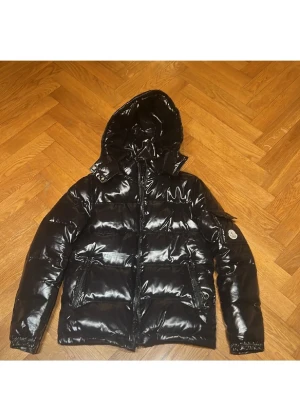 Svart glansig dunjacka från Moncler - Snygg svart dunjacka från Moncler med glansig finish och huva. Jackan har dragkedja framtill, två stora fickor och en Moncler-logga på ärmen. Perfekt för kalla dagar när du vill hålla stilen och värmen.