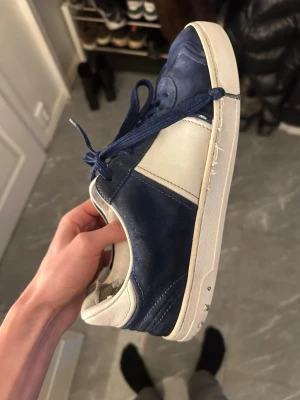 Blåvita sneakers med snörning - Snygga sneakers i blått och vitt med klassisk design och rund tå. Skorna har snörning och en vit platt sula. Materialet ser ut att vara skinn eller syntet, vilket ger en stilren look som passar till många outfits.