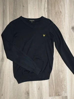 Svart finstickad tröja Lyle & Scott - Svart finstickad tröja från Lyle & Scott med V-ringning och klassisk gul logga på bröstet. Tröjan har långa ärmar och ribbade muddar vid ärmslut och nederkant. Perfekt för en stilren och enkel look.