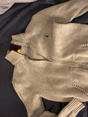 Beige stickad tröja Polo Ralph Lauren - Beige stickad tröja från Polo Ralph Lauren med dragkedja framtill och hög krage. Tröjan har ribbstickade muddar och en detalj med mörkröda och svarta ränder på kragen. Perfekt för lager-på-lager och snygg till jeans. Förutom den lilla fläcken är tröjan i bra skick.
