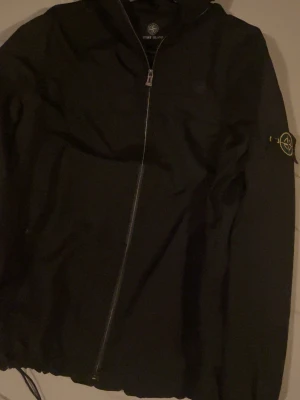 Svart vindjacka från Stone Island - Snygg svart vindjacka från Stone Island med ikonisk patch på ärmen. Jackan har dragkedja framtill, huva och en minimalistisk design. Tillverkad i lätt och slitstark polyester, perfekt för blåsiga dagar.