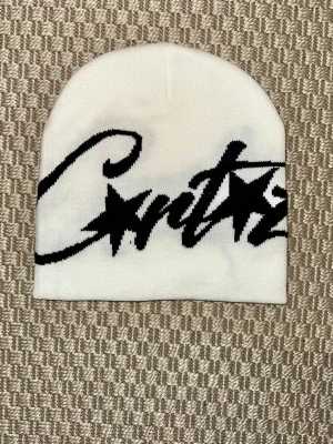 Corteiz mössa vit med svart logga  - Säljer en vit mössa med svart graffiti-inspirerad text och stjärndetaljer. Mössan är mjuk och stretchig, perfekt för att hålla värmen.