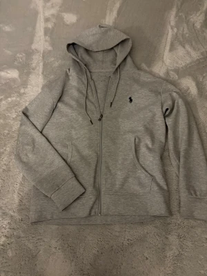 Grå hoodie från Polo Ralph Lauren - Hör av innan köp! Snygg grå zip-hoodie från Polo Ralph Lauren med klassisk huva och dragsko. Broderad logga på bröstet och två fickor framtill. Tillverkad i mjuk bomullsblandning, perfekt för en avslappnad stil.