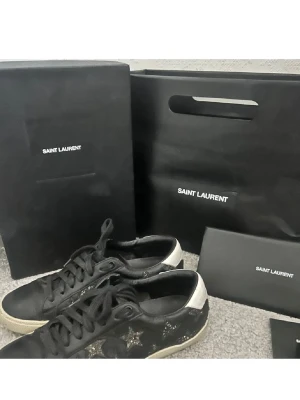 Svarta Saint Laurent sneakers med stjärnor - Svarta sneakers från Saint Laurent med glittriga stjärnor på sidorna och vita detaljer vid hälen. Skorna har snörning och är tillverkade i skinn, vilket ger en lyxig känsla. Perfekta för dig som vill sticka ut med en unik och trendig look.