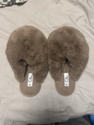 Bruna fluffiga tofflor från Chlox - Mysiga bruna tofflor från Chlox med supermjuk och fluffig pälsimitation. Tofflorna har öppen tå och platt sula, perfekta för att glida runt hemma i. Snygg och enkel design som känns lyxig och bekväm.