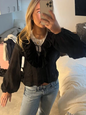 Svart blus med volang och snörning - Snygg svart blus med volangdetaljer runt halsen och snörning framtill. Blusen har långa, puffiga ärmar med resår vid handlederna och är tillverkad i ett lätt, något transparent tyg. Perfekt att styla med jeans för en trendig look.