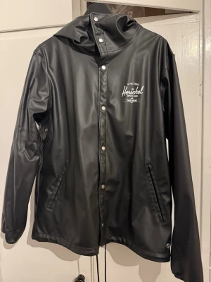 Svart regnjacka från Herschel - Snygg svart regnjacka från Herschel med huva och tryckknappar framtill. Jackan har två fickor, dragsko nedtill och logga på bröstet. Tillverkad i vattenavvisande syntetmaterial och har lång ärm. Perfekt för regniga dagar.