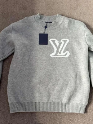 Grå stickad tröja  - Snygg grå stickad tmed stort vitt logo på bröstet. Tröjan har rund halsringning och ribbade muddar vid ärmslut och nederkant. 