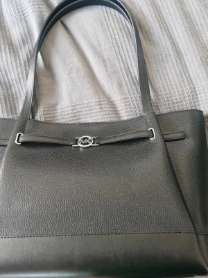 Svart handväska från Michael Kors - Stilren svart handväska från Michael Kors med silverfärgad logodetalj framtill. Väskan har dubbla axelband och är tillverkad i skinnimitation med struktur. Insidan är ljus och rymlig, perfekt för vardagens alla prylar. Finns ett litet lite märke på insidan, se bild. Använd 1 gång.