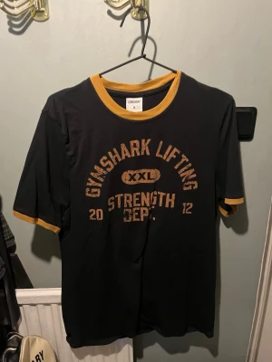 Svart Gymshark t-shirt med gula detaljer - Svart t-shirt från Gymshark med gula muddar vid ärmslut och hals. Tryck på bröstet med texten 'GYMSHARK LIFTING STRENGTH DEPT. 2012' i gul vintage-stil. Perfekt för dig som gillar sportig och avslappnad stil.