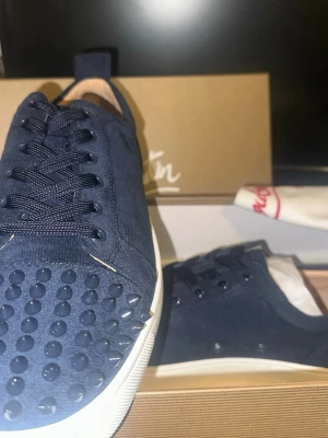 Mörkblå sneakers med nitar från Louboutin - Snygga mörkblå sneakers från Christian Louboutin i mocka med coola svarta nitar på tån. Skorna har snörning och vit platt sula. Perfekta för dig som vill sticka ut med en lyxig och edgy stil.