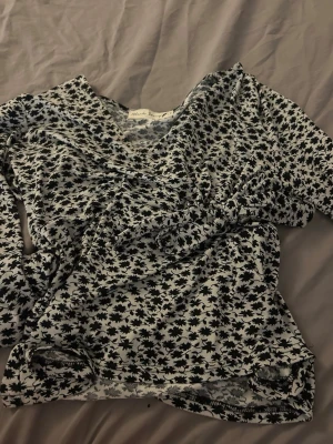 Svartvit blommig omlottopp Black Story - Trendig omlottopp från Black Story i svartvitt blommigt mönster. Toppen har v-ringning och långa ärmar, samt en snygg omlottdetalj framtill.