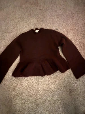 Vinröd stickad volangtröja - Säljer en vinröd stickad tröja med volangdetalj nedtill och rund hals. Tröjan har långa ärmar och en något lös passform. Perfekt för dig som gillar en unik twist på klassiska stickade plagg.