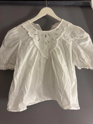 Vit blus med spetsdetaljer Gina Tricot - Söt vit blus från Gina Tricot med puffiga korta ärmar och vacker spetsdetalj vid halsringningen. Blusen har broderade mönster och en luftig, lätt struktur. Perfekt för en trendig och romantisk look.