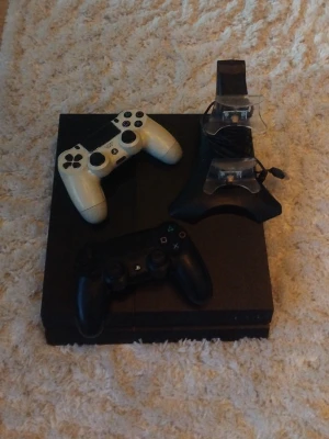 Playstation 4 med två handkontroller - Säljer en svart Playstation 4 med en svart och en vit DualShock 4-handkontroll samt en laddstation för kontrollerna. Konsolen har en stilren, fyrkantig design och är tillverkad i plast. Perfekt för gamingkvällar med vänner.