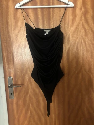 Svart draperad body från H&M - Snygg svart body från H&M med tunna axelband och draperad framsida. Tillverkad i mjukt stretchigt material som sitter skönt mot kroppen. Perfekt för att skapa en trendig och stilren look. Helt ny 