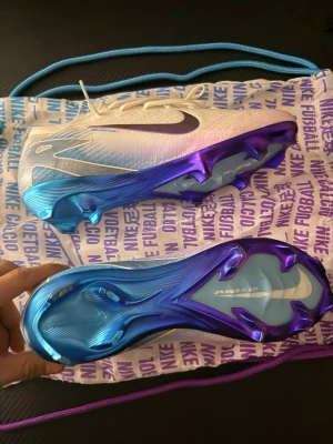 Nike Mercurial fotbollsskor blå/vit - Nike Mercurial fotbollsskor i vitt med blå och lila detaljer. Skorna har en snygg gradient från vit till blå, lila sula och klassisk Nike-logga på sidan. Ovandelen är i syntetmaterial och skon har snörning för bra passform. Perfekta för dig som vill sticka ut på planen. Storlek 42.5