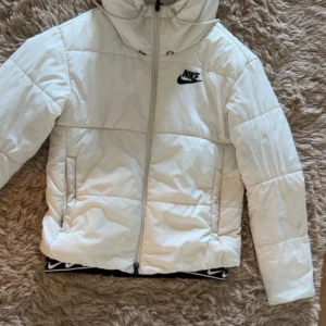 Vit pufferjacka från Nike med huva - Säljer en snygg vit pufferjacka från Nike i storlek S. Jackan har huva, dragkedja framtill och svart Nike-logga på bröstet. Perfekt för kyliga dagar och har en clean, sportig look. Jackan är vadderad och har två fickor på sidorna.