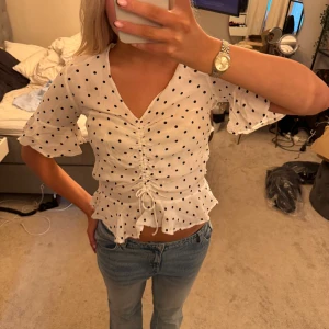 Vit prickig blus med snörning - Supersöt vit blus med svarta prickar och v-ringning. Toppen har volangiga ärmar, snörning framtill och lätt croppad passform. Materialet känns lätt och luftigt, perfekt för varmare dagar. Snygg detalj med volangkant nertill och knytning i midjan.