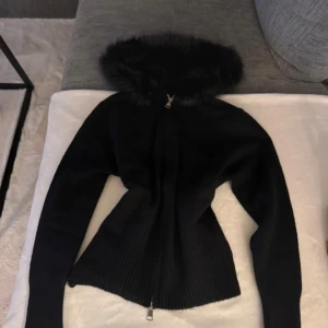 Alessa liknande jacka - Svart stickad jacka med dragkedja framtill och stor huva med fuskpäls. Jackan har ribbade muddar och en figurnära passform som ger en snygg siluett. Perfekt för kyliga dagar när du vill vara både varm och stilren.