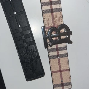 120 cm bälte från burrbery  - Ett snyggt bälte från Burberry – ett i klassiskt beige rutigt mönster med silverfärgat BB-spänne och ett i svart med diskret rutig struktur och präglad ryttare. Båda är i skinn och har bred design som sticker ut till jeans eller kostymbyxor. Man kan byta sida på bältet och få en svart färg eller beige brun färg.