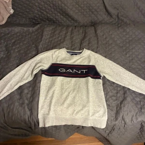 Grå sweatshirt från GANT med logga - Säljer en stilren grå sweatshirt från GANT med klassisk logga över bröstet i vitt på en marinblå panel med röda detaljer. Tröjan har rund halsringning och långa ärmar. Perfekt för en chill och snygg look.