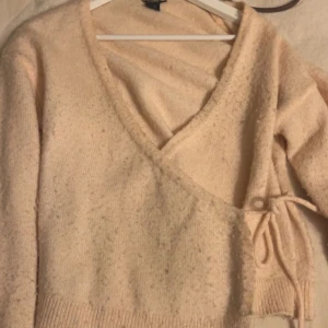 Beige omlott kofta med knyt - Supermysig beige omlott kofta med knyt i sidan och v-ringad hals. Stickad i mjukt material och har långa ärmar samt ribbade muddar. Perfekt för lager-på-lager och chill dagar.