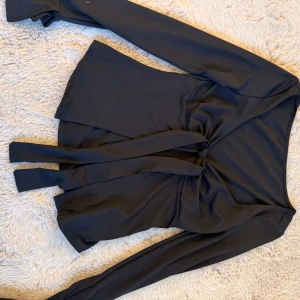 Svart omlottopp med knytband - Snygg svart omlottopp med långa ärmar och knytband i midjan. Toppen har en djup v-ringning och är tillverkad i ett mjukt, stretchigt material som sitter skönt på kroppen. Perfekt för dig som gillar stilrena och trendiga plagg.