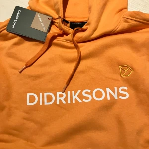 Orange hoodie från Didriksons i bomull - Snygg orange hoodie från Didriksons i ekologisk bomull. Tröjan har huva med snörning, broderad logga på ärmen och bröstet samt vit printad text framtill. Ribbstickade muddar och relaxed passform. Perfekt för chill dagar. Nypris 900,- 