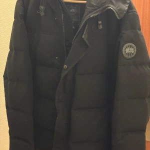 Svart dunjacka från Canada Goose - Säljer en svart dunjacka från Canada Goose med huva och dragkedja. Jackan har quiltad design, logotyp på ärmen och praktiska fickor. Perfekt för kalla dagar och har en clean, stilren look som funkar till det mesta. pris kan diskuteras vid snabb affär dragkedjan till yttefickorna har gått sönder dom medföljer också om man önskar de