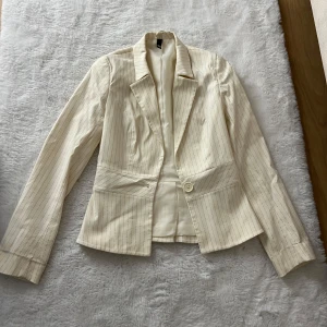 Randig beige kostym med kavaj och byxa - Snygg tvådelad kostym från i beige med tunna vertikala ränder. Kavajen har klassisk krage, en knapp och figursydd passform. Byxorna är utsvängda med låg midja . Perfekt för dig som vill sticka ut med en stilren och trendig look. Kavajen säljs separat för 299 samt byxorna. Komplett 500kr
