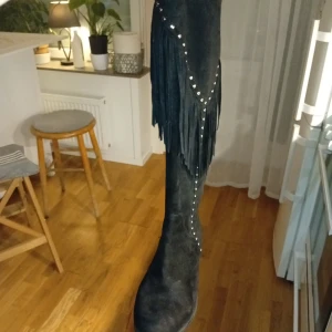 Svarta mockaimiterade stövlar med fransar och nitar - Snygga svarta stövlar med coola fransdetaljer och silverfärgade nitar längs skaftet. Stövlarna har platt sula, rund tå och dragkedja på insidan. Perfekta för dig som vill sticka ut med en westerninspirerad look.
