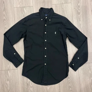 Svart skjorta från Ralph Lauren - Svart långärmad skjorta från Ralph Lauren i slim fit-modell. Skjortan har klassisk button down-krage, vita knappar och den ikoniska broderade loggan på bröstet. Tillverkad i mjuk bomull, perfekt för en clean och stilren look.