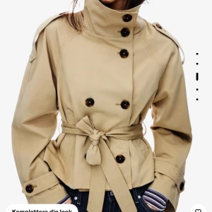 Beige trenchcoat med bälte - Stilren beige trenchcoat med hög krage, dubbelknäppning och knytskärp i midjan. Jackan har axelklaffar, stora knappar och klassiska detaljer som ger en snygg siluett. Perfekt för dig som vill ha en tidlös och trendig look.