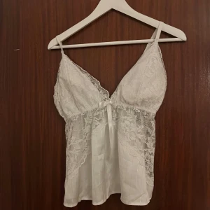 Vit spetsig linnetopp med rosett - Supersöt vit linnetopp med tunna axelband, spetsdetaljer och liten rosett framtill. Toppen har v-ringning och är tillverkad i glansigt material med blommig spets och lätt genomskinliga partier. Perfekt för dig som gillar romantisk och feminin stil.