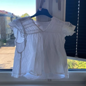 Vit blus med volang från Gina Tricot - Supersöt vit blus från Gina Tricot med volangärmar och v-ringning. Blusen har broderade detaljer upptill och är tillverkad i tunn, luftig bomull. Perfekt för varma dagar och ger en romantisk vibe till din outfit.