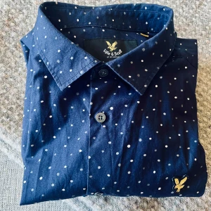 Mörkblå prickig skjorta Lyle & Scott - Stl M. Snygg mörkblå skjorta från Lyle & Scott med vita prickar och klassisk krage. Skjortan har långa ärmar, knappar framtill och den ikoniska guldfågeln broderad på bröstet. Perfekt för dig som gillar stilrena detaljer och mönster.