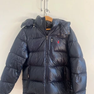 Svart pufferjacka från Polo Ralph Lauren - Pufferjacka från Polo Ralph Lauren med glansig finish och röd logga på bröstet. Jackan har huva, flera dragkedjefickor och quiltad design. Perfekt för kalla dagar och har en riktigt chill vibe. Passar dig som gillar streetstyle och vill hålla värmen. Använd en vinter samt Kvitto finns. Priset är inte hugget i sten kom med förslag, tveka inte att att fråga frågor:) nypris 6099kr (mörkblå)