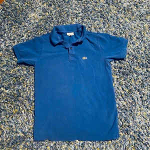 Blå pikétröja från Lacoste - Klassisk blå pikétröja från Lacoste med korta ärmar och ikoniska krokodilloggan broderad på bröstet. Tröjan har krage, knappar framtill och är tillverkad i bomull för en skön och luftig känsla. Perfekt för en clean och sportig stil.