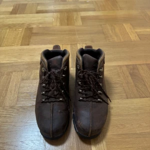 Bruna Timberland kängor i skinn - Säljer ett par bruna kängor från Timberland i robust skinn med snörning och grov sula. Skorna har rund tå och detaljer i mörkbrunt och beige, perfekta för dig som gillar outdoor-vibe och klassisk stil. Perfekt för vintern! 