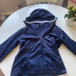 Vindjacka polo Ralph lauren barnstorlek XL passar S - Snygg marinblå vindjacka från Polo Ralph Lauren i storlek XL. Jackan har huva, dragkedja framtill, två fickor med dragkedja och ribbade muddar med vita ränder. Perfekt för blåsiga dagar och stilren look.