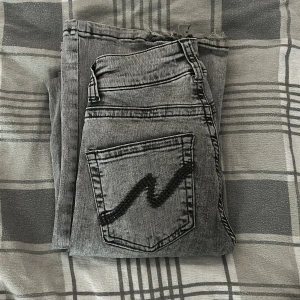 Grå jeans från Nelly med rå kant - Snygga grå jeans från Nelly med tvättad look och svarta detaljer på bakfickan. Modellen har hög midja, raka ben och rå kant nedtill för en cool vibe. Dubbelknäppning framtill och klassiska fem fickor. Perfekt för dig som gillar streetstyle.